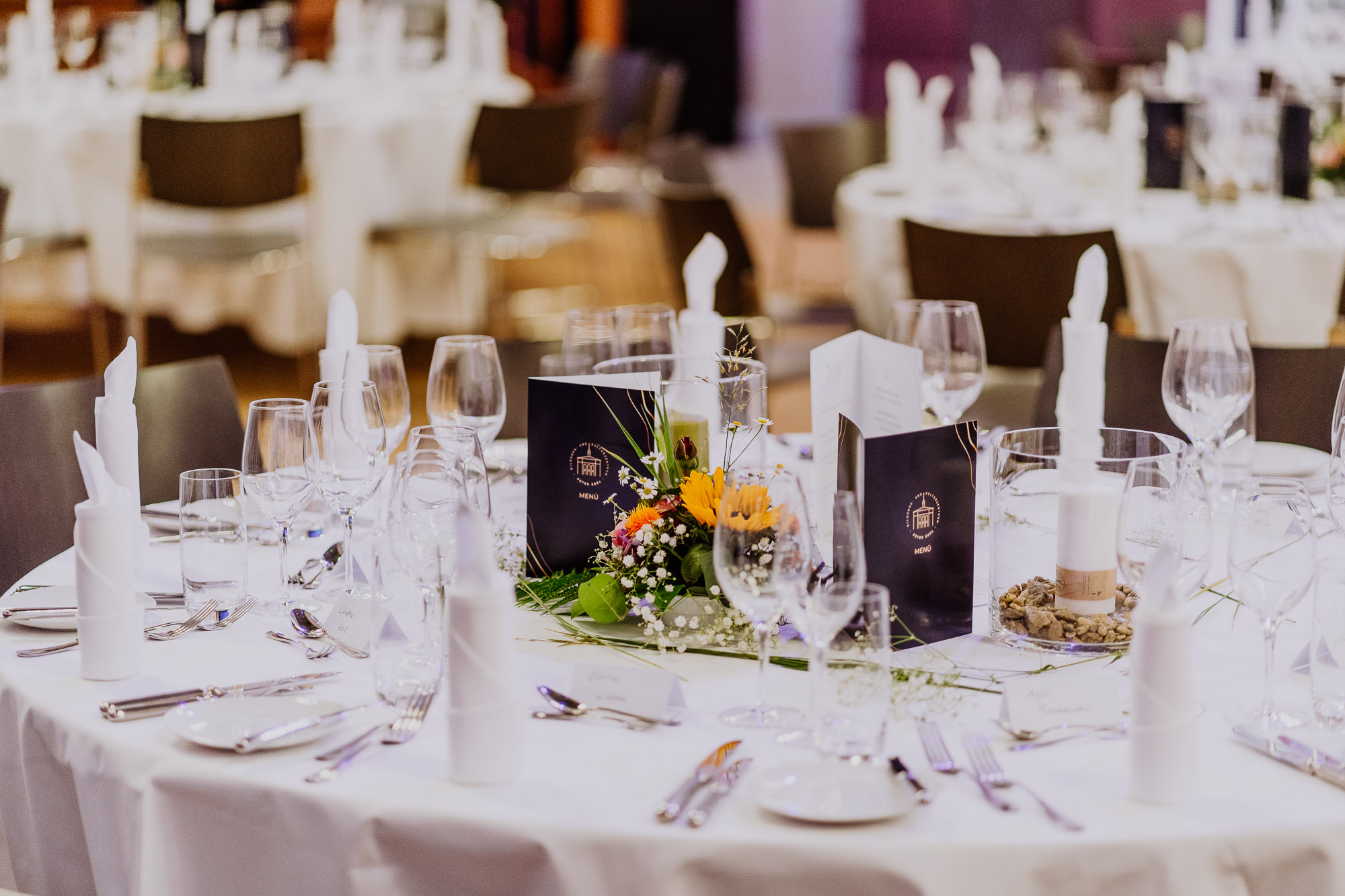 Ein elegant eingedeckter Tisch mit einer weißen Tischdecke, Blumen, Speisekarten im PETER EDEL-Stil und Besteck