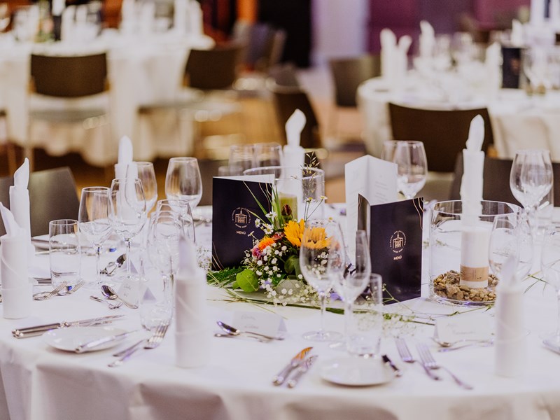 Ein elegant eingedeckter Tisch mit einer weißen Tischdecke, Blumen, Speisekarten im PETER EDEL-Stil und Besteck
